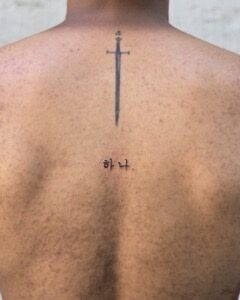 Korean lettering tattoo 하나. with a small fine-line sword on the upper back, Seoul