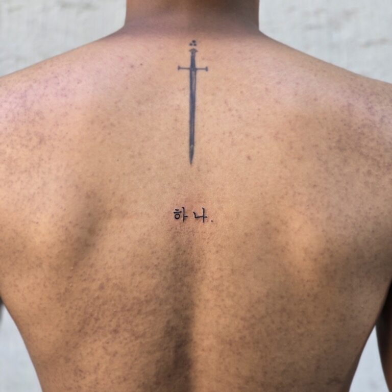 Korean lettering tattoo 하나. with a small fine-line sword on the upper back, Seoul
